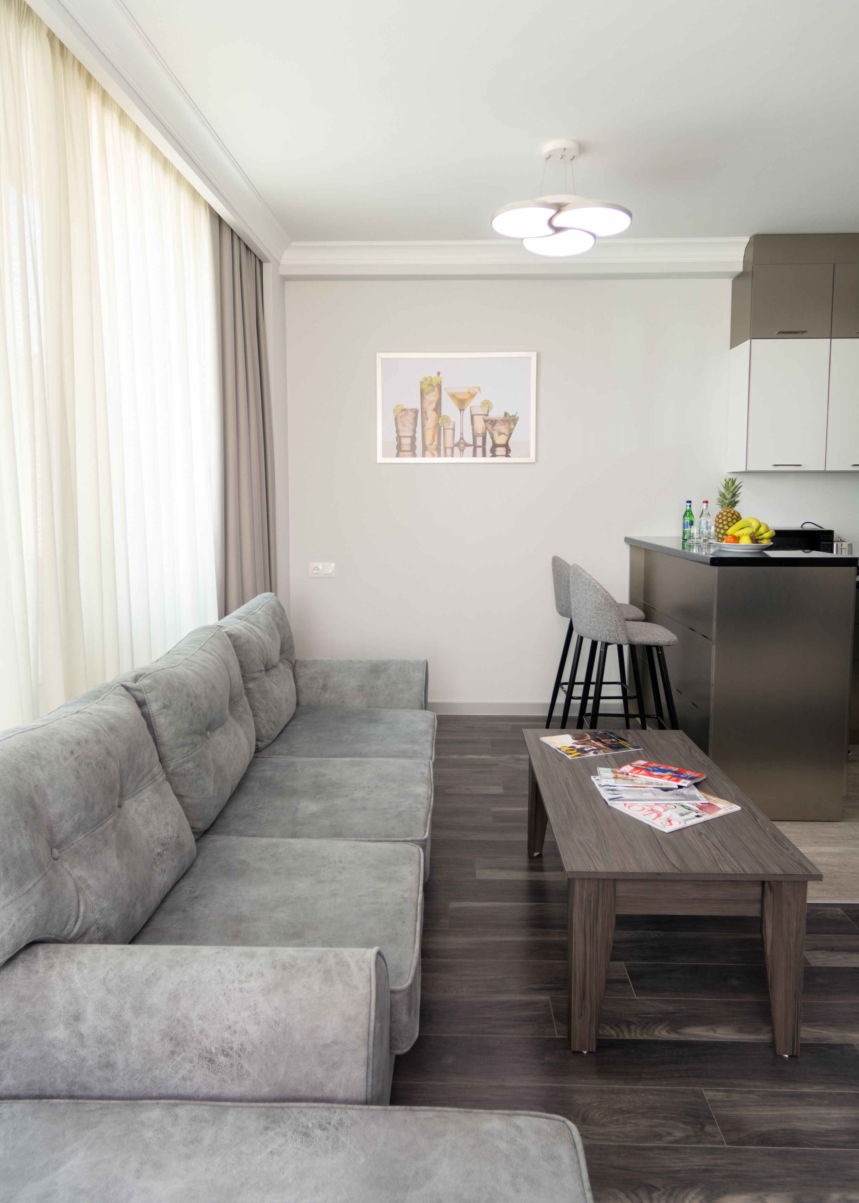A modern living room with a grey sectional sofa, a wooden coffee table with magazines, a small bar area with stools, and a kitchen with cabinets and a refrigerator.