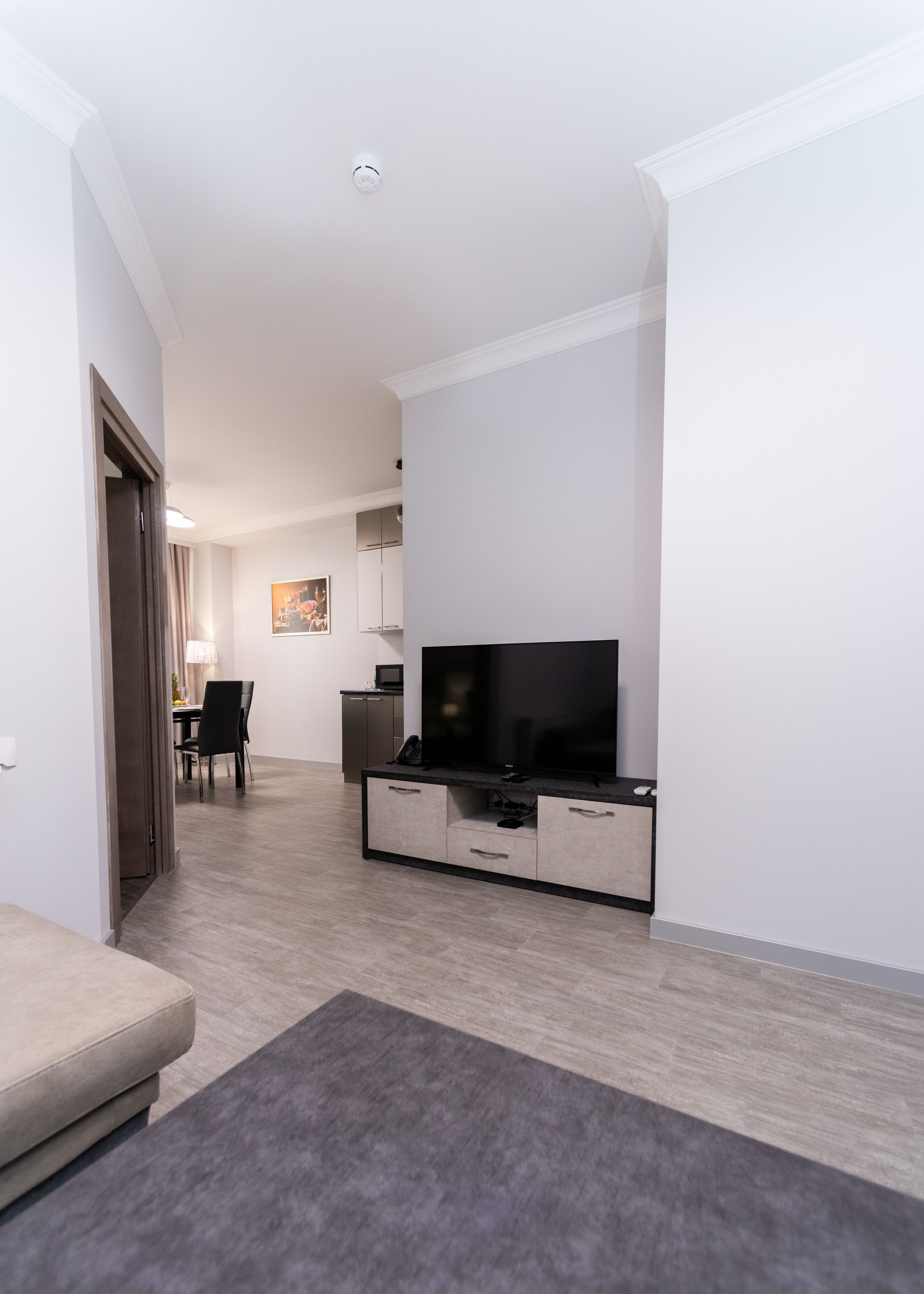 Een moderne woonkamer met een flatscreen televisie, een eethoek met een tafel en stoelen, en een kitchenette met kastjes en een magnetron.