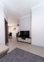 Een moderne woonkamer met een flatscreen televisie, een eethoek met een tafel en stoelen, en een kitchenette met kastjes en een magnetron.