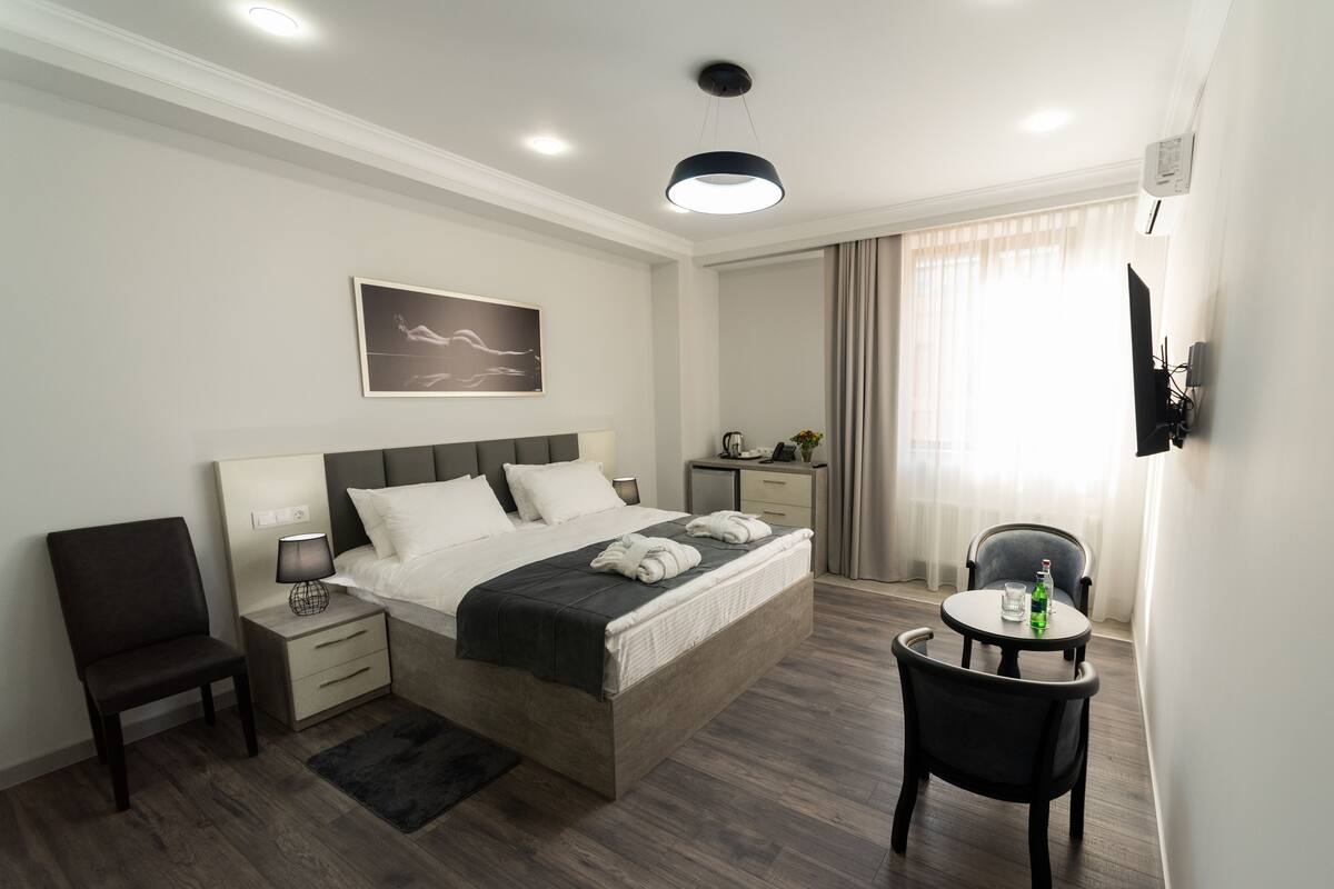 A modern hotel room with a large bed, bedside tables, a desk, a chair, and a TV mounted on the wall.