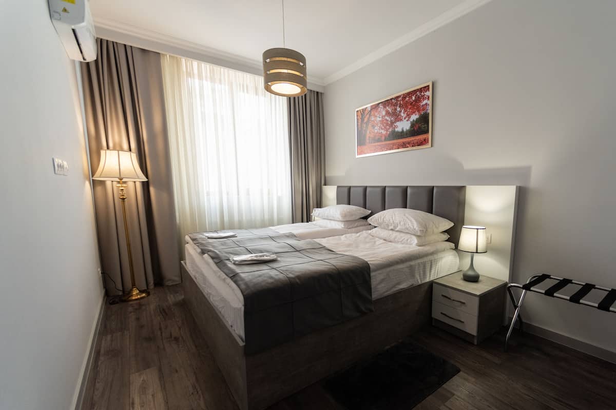A modern hotel room with a large bed, bedside tables, a standing lamp, a black and white striped towel rack, and a window with sheer curtains.