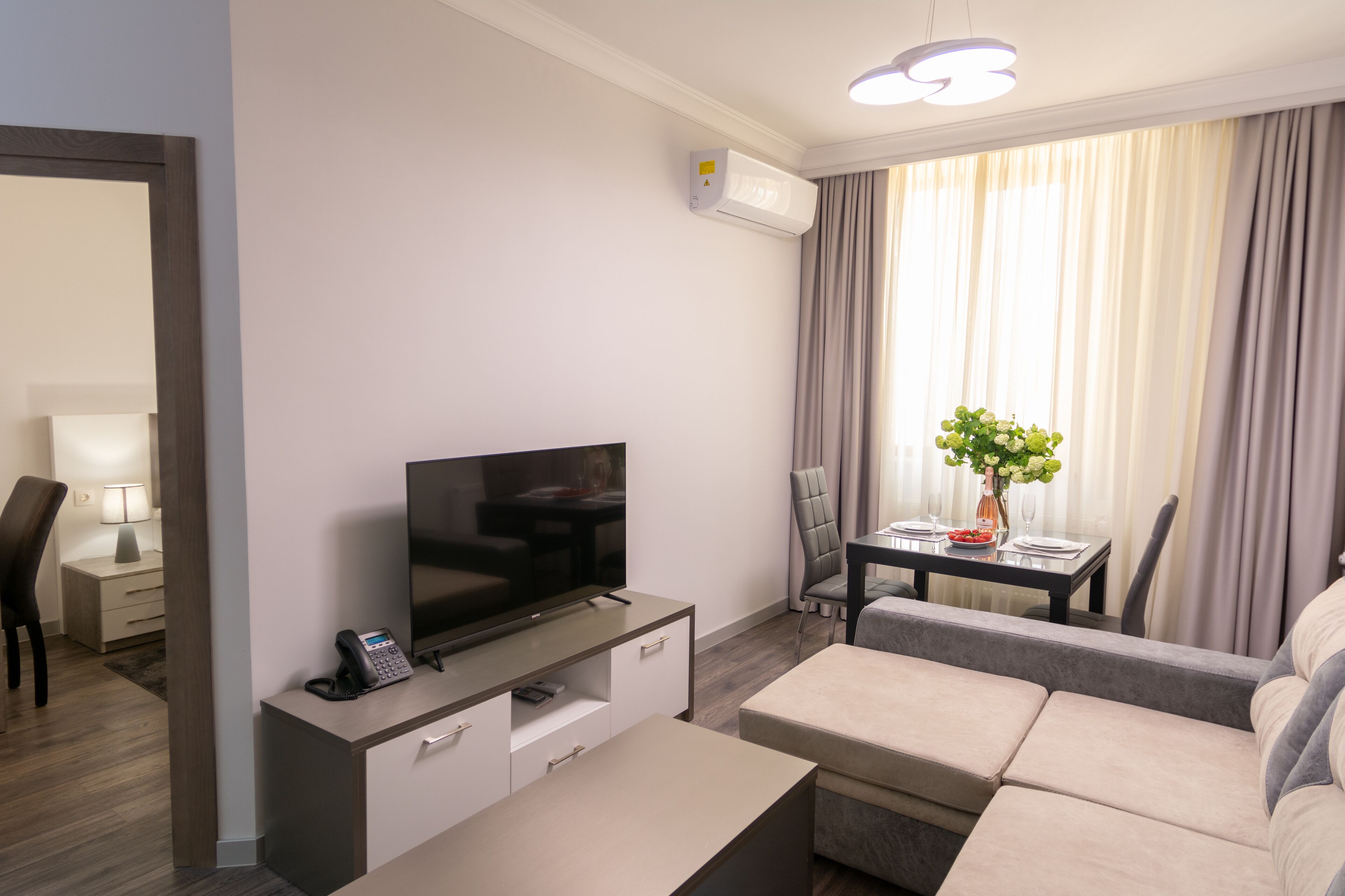 A modern hotel room with a flat-screen TV on a wooden console, a sofa, a dining table with chairs, and a large window with curtains.