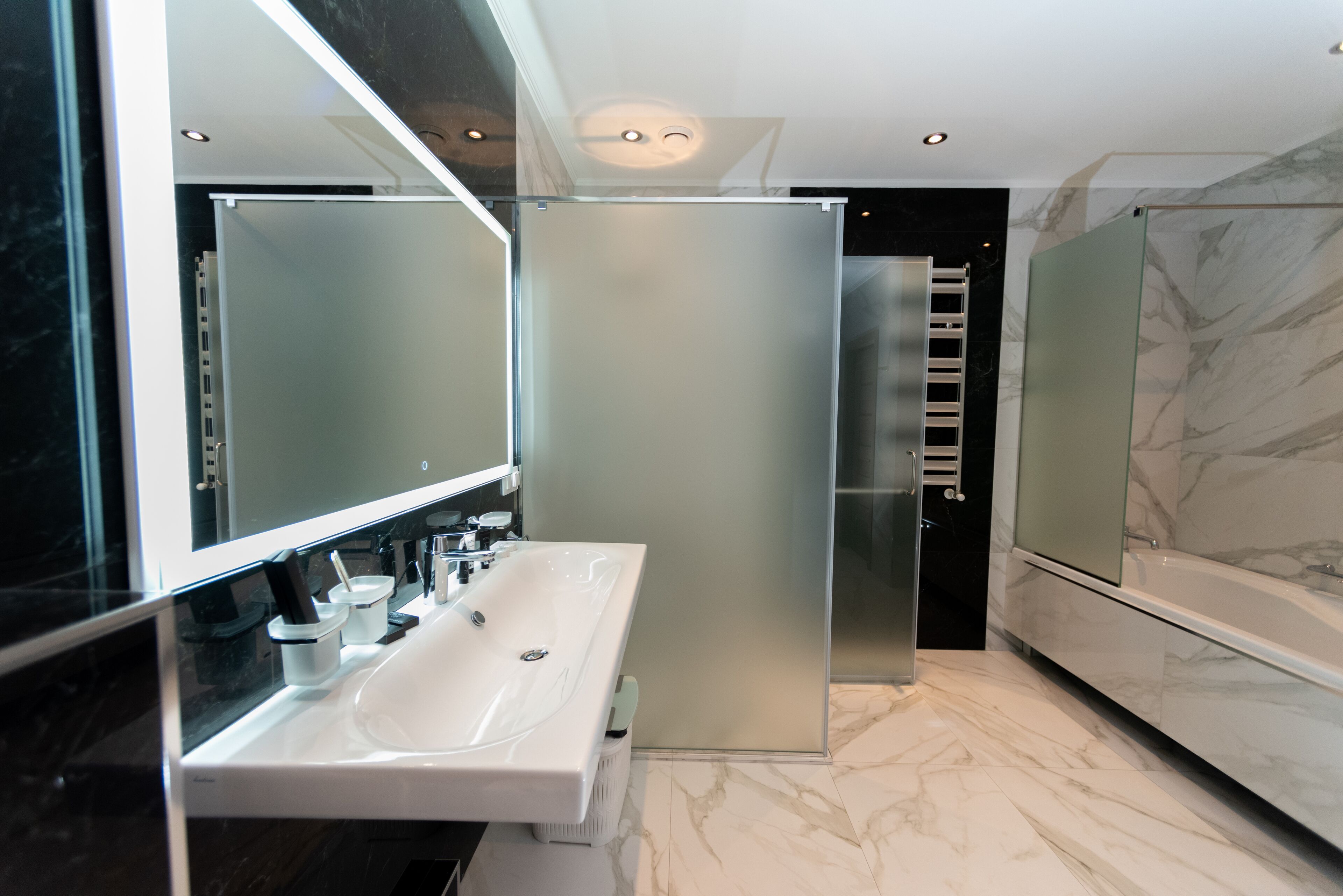 A modern bathroom with a large mirror, a white sink, a glass shower enclosure, and a marble floor.