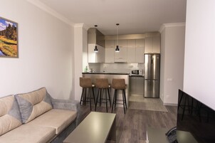 Een moderne woonkamer met een bank, een eettafel en barkrukken. In de achtergrond is een keuken met een koelkast en een bar te zien.