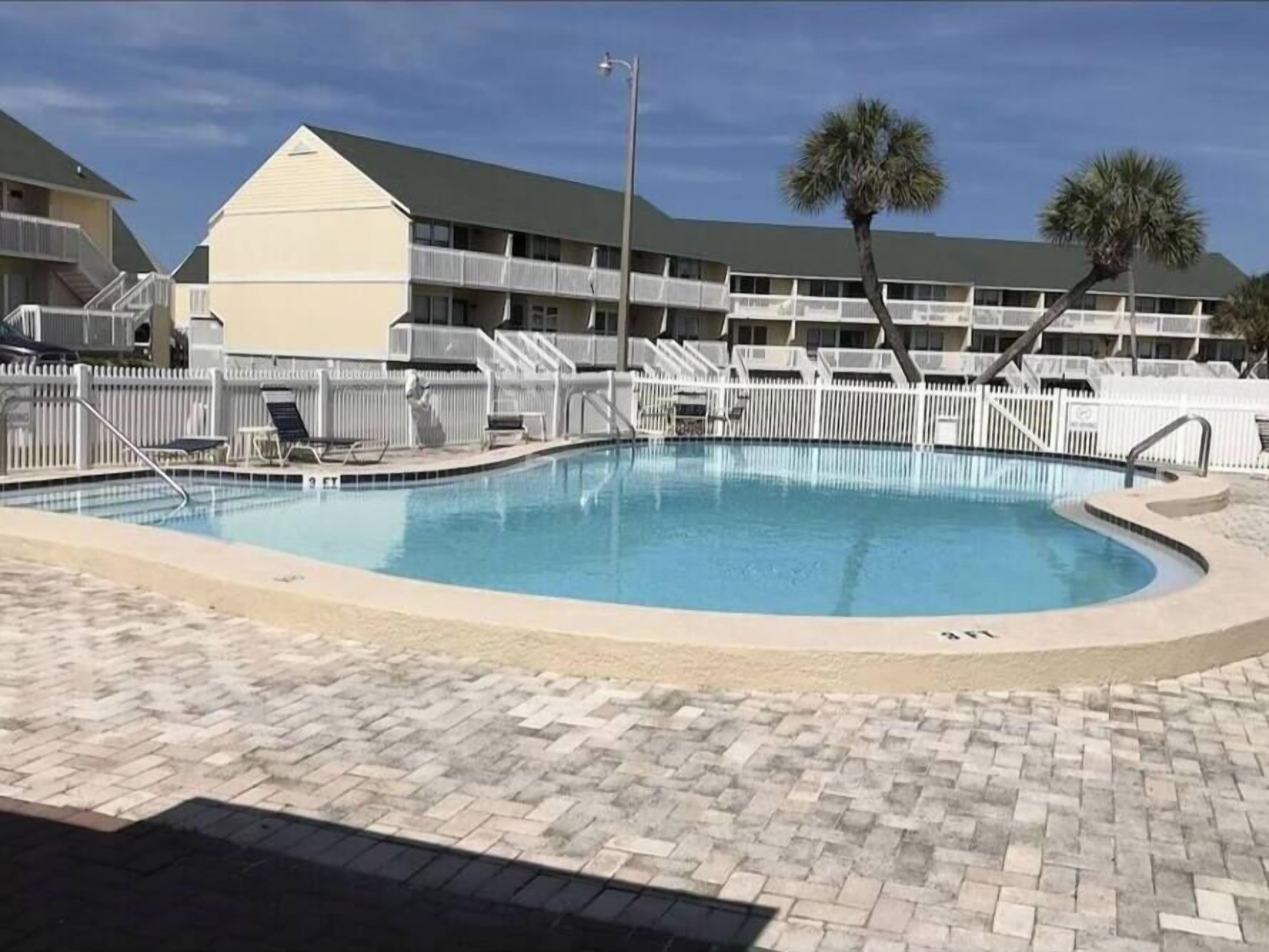 A swimming pool with a brick deck, surrounded by a white fence and palm trees.