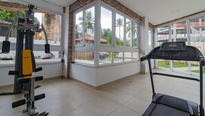 A gym with a treadmill, weight machines, and large windows offering a view of palm trees and buildings. - Apart Porto de Galinhas para 4 pessoas (Ipojuca)