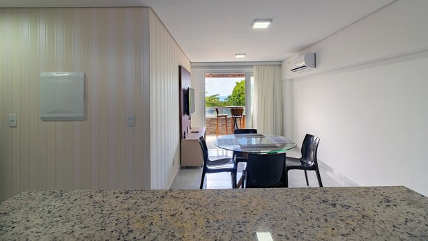 A modern room with a granite countertop, a dining table with chairs, a TV mounted on the wall, and a view of the outdoors through a window. - Apart Porto de Galinhas para 4 pessoas (Ipojuca)