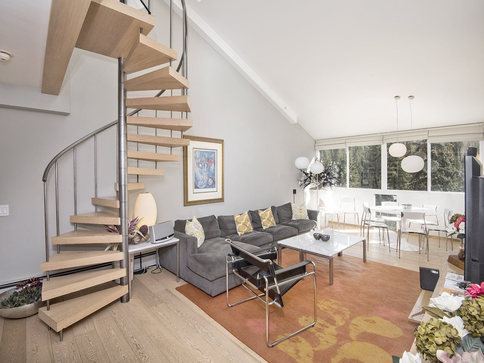 A modern living room with a spiral staircase, grey sofa, coffee table, and a dining area with white chairs.