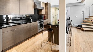 A modern kitchen with wooden cabinets, a black countertop, and stainless steel appliances. There is a dining area with a bar stool and a staircase leading to another level. - Lionshead Center (Vail)