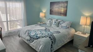 A bedroom with a bed, two bedside lamps, a ceiling fan, a window with sheer curtains, and a beach-themed wall art. - Siesta Key Condo- walk to beautiful beaches, ride a free trolley around the key  (Sarasota)