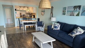 A living room with a sofa, coffee table, and ottoman. There is a dining area with a table and chairs. The room has wooden flooring and light blue walls. - Siesta Key Condo- walk to beautiful beaches, ride a free trolley around the key  (Sarasota)