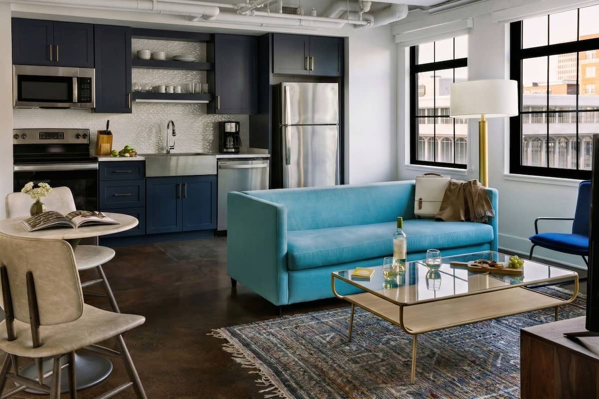 A modern kitchen with dark cabinets, a stainless steel refrigerator, and a microwave. There is a blue sofa, a glass coffee table, and a dining area with chairs.