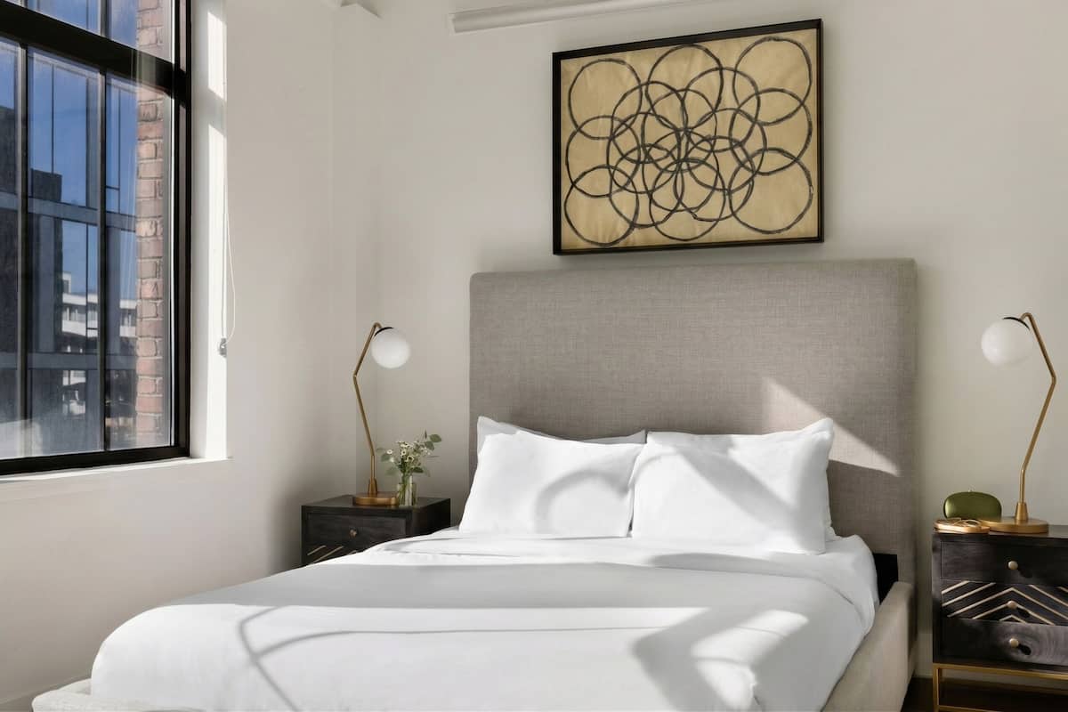 A neatly made bed with white linens, a grey upholstered headboard, and a framed abstract artwork above it.