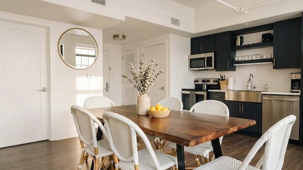 A modern kitchen with dark cabinets, a wooden dining table, wicker chairs, and a round mirror.