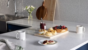 A kitchen counter with a sink, a wooden cutting board, a bowl of berries, a jar of honey, a plate with scones, and two white mugs.