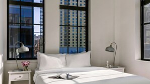 A hotel room with a large window, a bed, a nightstand with a lamp, and a vase with flowers.