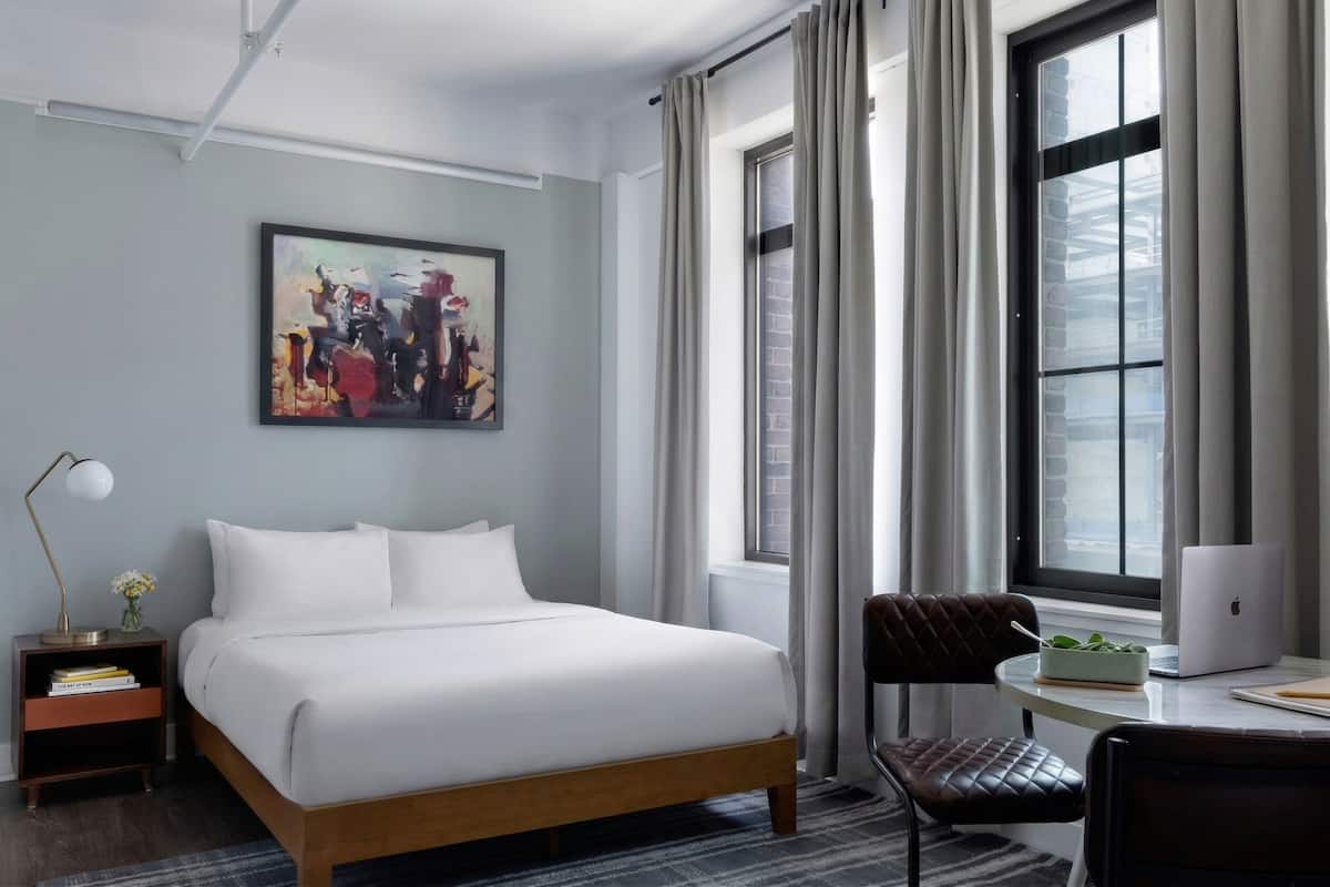 A hotel room with a bed, a desk with a laptop, a chair, a nightstand with a lamp, and a painting on the wall.