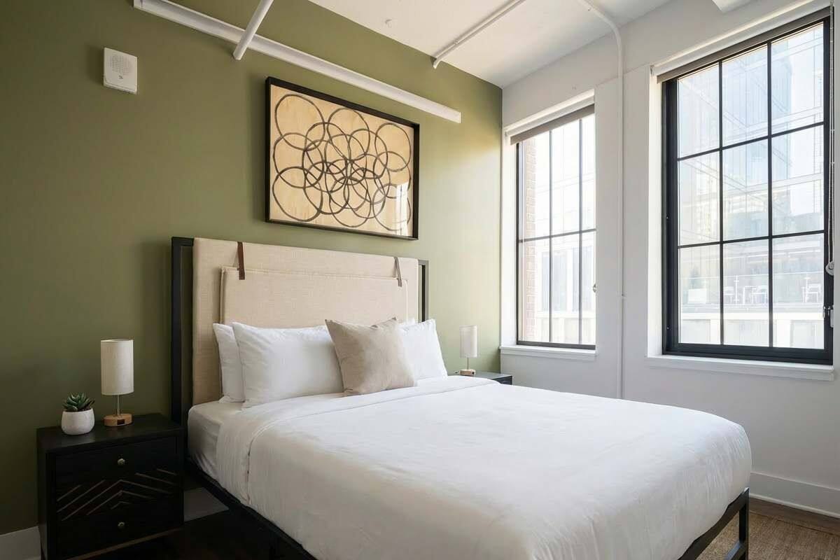 A neatly made bed in a room with a large window, a wall art piece, and a nightstand with a lamp.