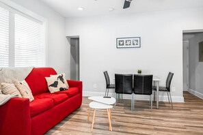 A modern living room with a red sofa, a small white table, and black chairs. There is a framed picture on the wall and a window with blinds.