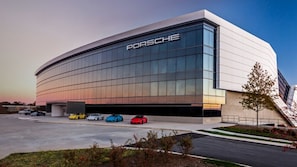 A modern building with a prominent Porsche logo, featuring a large parking area with several cars.