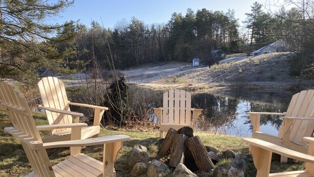 A lakeside setting with three wooden Adirondack chairs facing a small pond, surrounded by rocks and logs. - The Lodge House - Centrally Located Cheerful 5BR - Minutes to Gore! (North Creek)