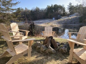 A lakeside setting with three wooden Adirondack chairs facing a small pond, surrounded by rocks and logs.