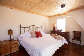 A bedroom with a wooden ceiling, a bed with white bedding, a nightstand with a lamp, a dresser, and a chair.