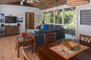 A spacious living area with a wooden dining table, a sofa, and a television mounted on the wall. - Blue Agave Villa- Dazzling Sunsets, Amazing Views, Pool, Generator A/C in Br's (Cruz Bay)