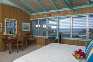 A wooden room with a bed, a desk, and a chair. There are windows with a view of the ocean. - Blue Agave Villa- Dazzling Sunsets, Amazing Views, Pool, Generator A/C in Br's (Cruz Bay)
