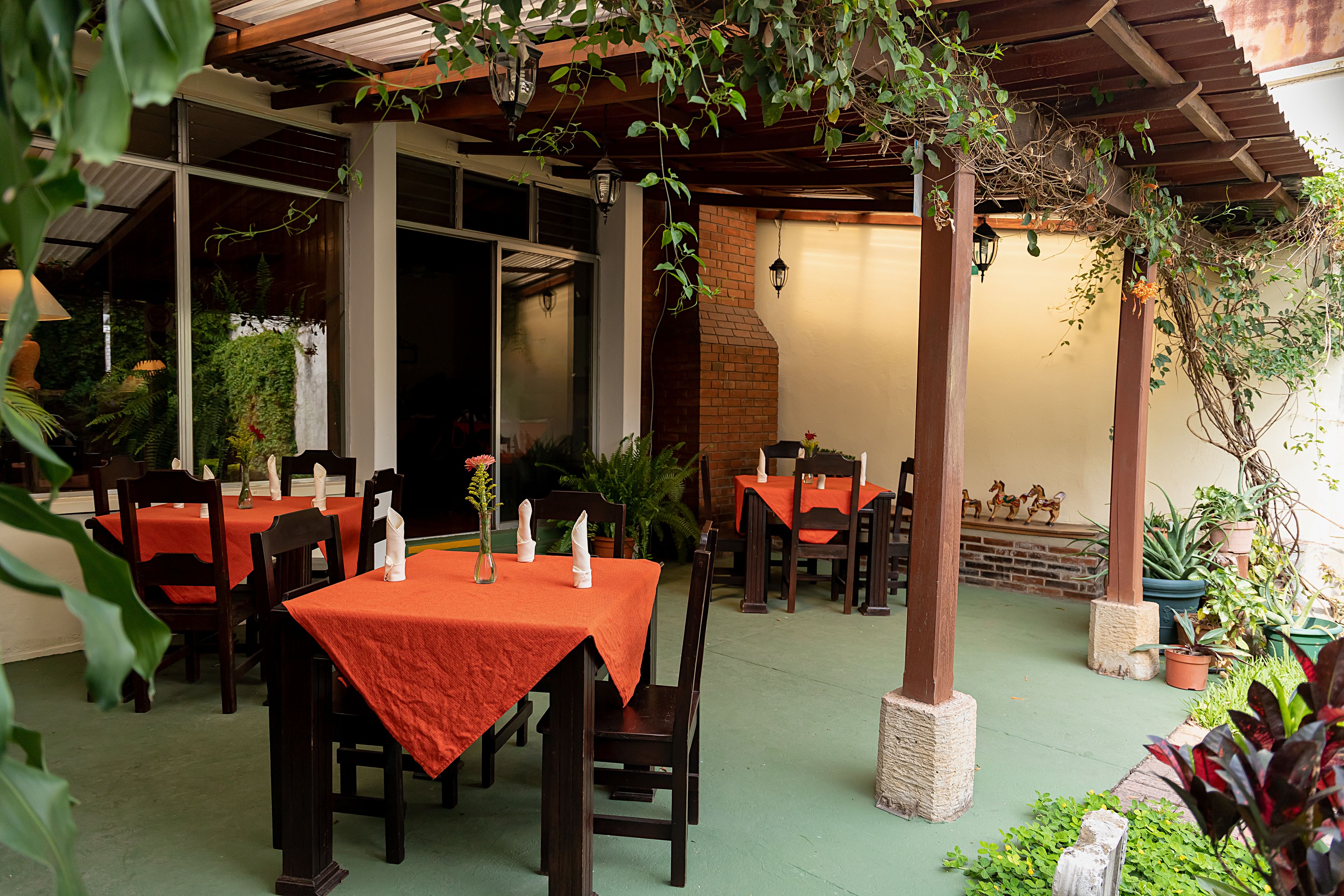 An outdoor dining area with tables and chairs, covered by a wooden pergola, and surrounded by potted plants.