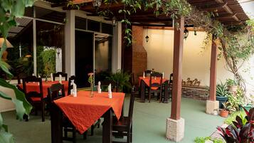 An outdoor dining area with tables and chairs, covered by a wooden pergola, and surrounded by potted plants.