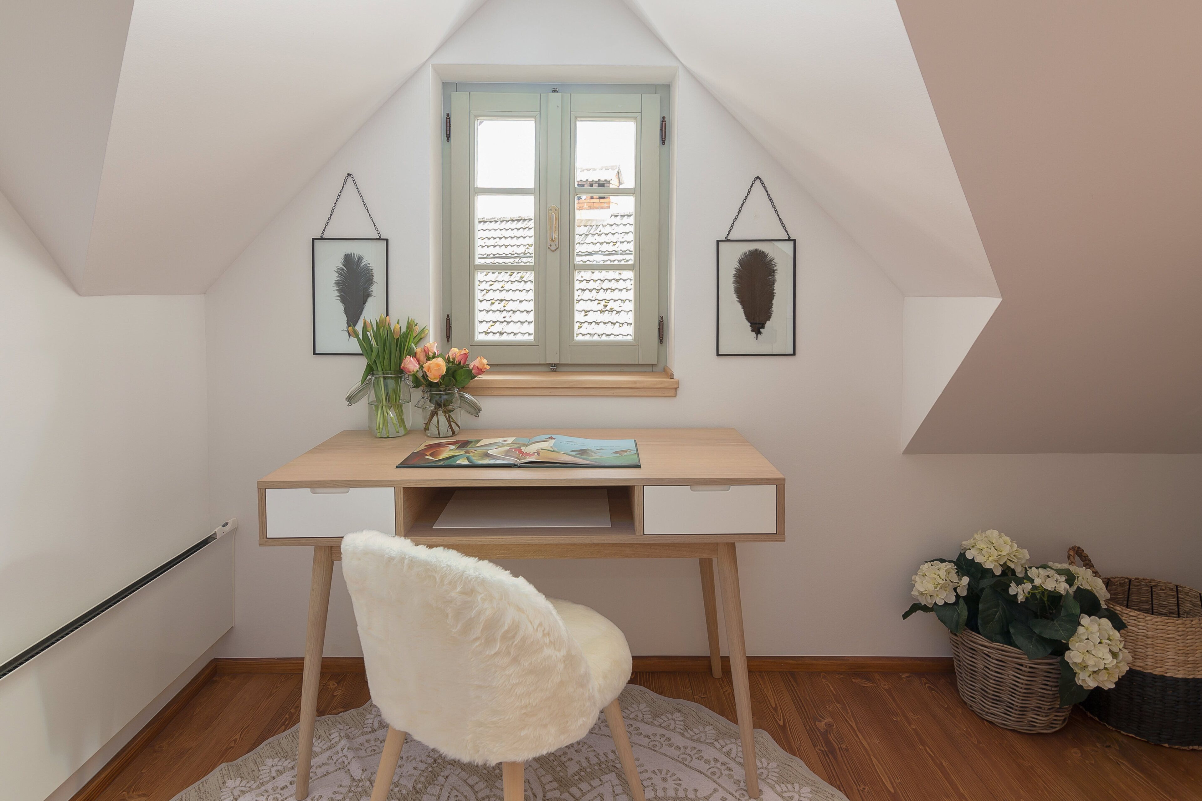 Une pièce avec un plafond en pente, un bureau en bois avec une chaise, une fenêtre donnant sur un beau paysage et deux tableaux accrochés au mur.