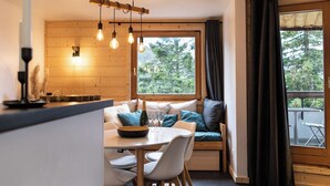 Un cuarto acogedor con detalles de madera, un comedor con mesa y sillas, un banco con cojines, y vistas a árboles a través de una ventana.