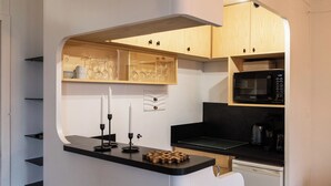 Una cocina compacta con microondas, cafetera y refrigerador pequeño. Cuenta con gabinetes de madera y una encimera negra.