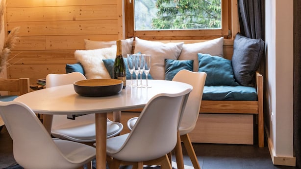 Una habitación acogedora con paredes de madera, una mesa de comedor con sillas, un sofá con cojines, un ventanal con vistas a la vegetación y una lámpara colgante.