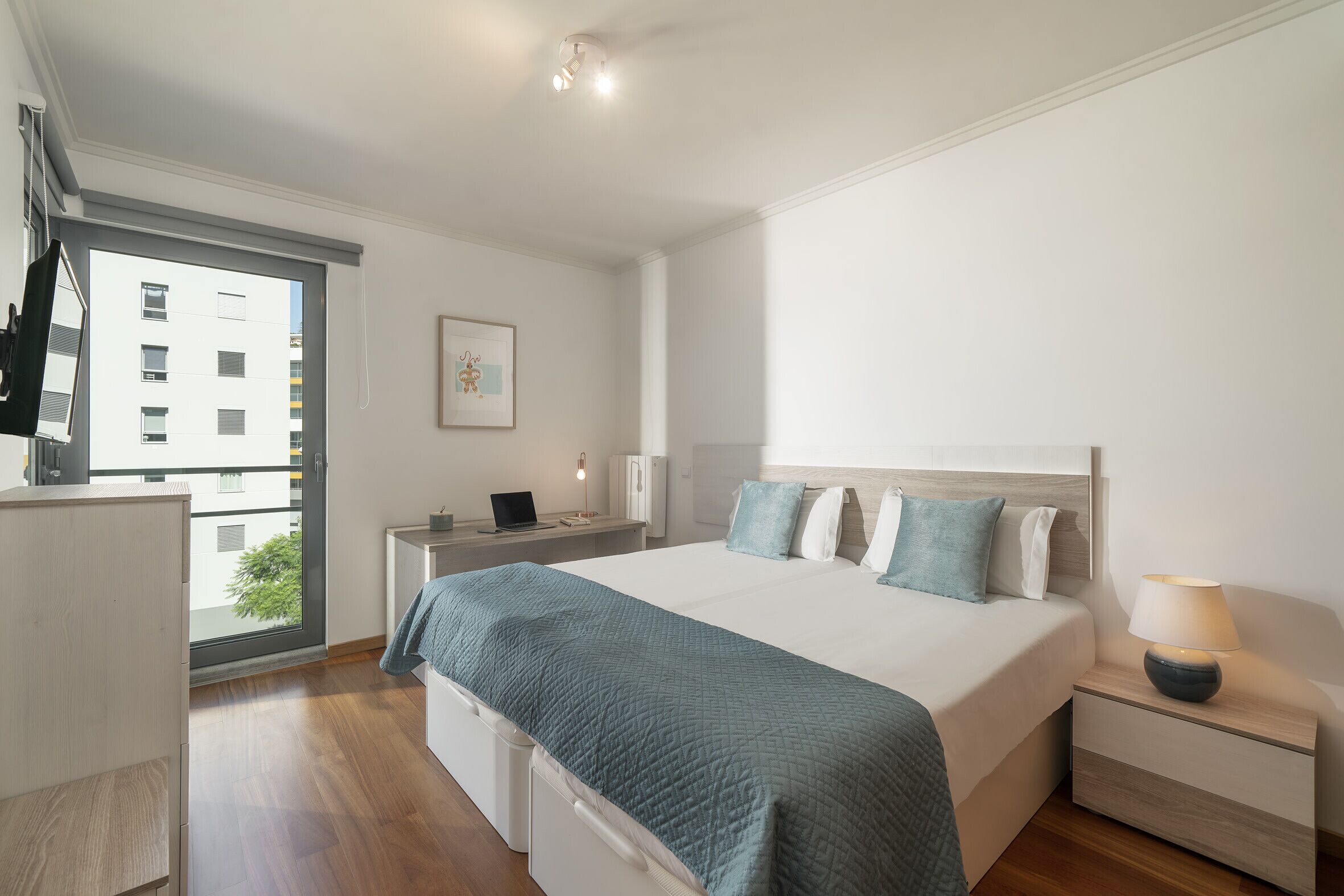 Un dormitorio moderno con una cama grande, mesitas de noche, un escritorio y una ventana con vistas a edificios.