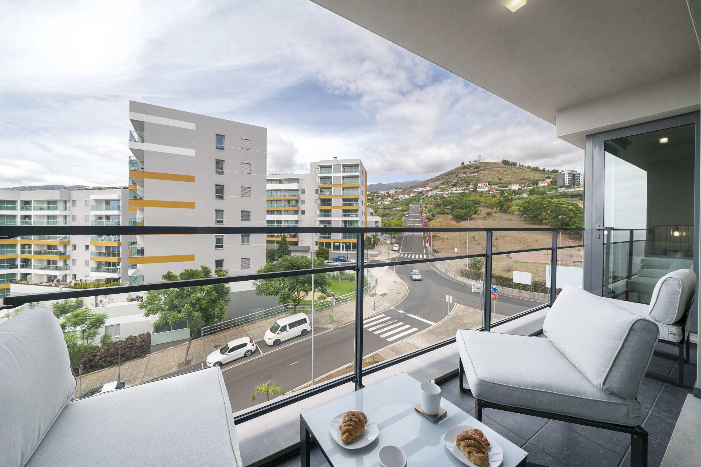 Een balkon met uitzicht op moderne gebouwen en een weg. Er staan twee witte banken en een klein tafeltje met een croissant en een kopje.