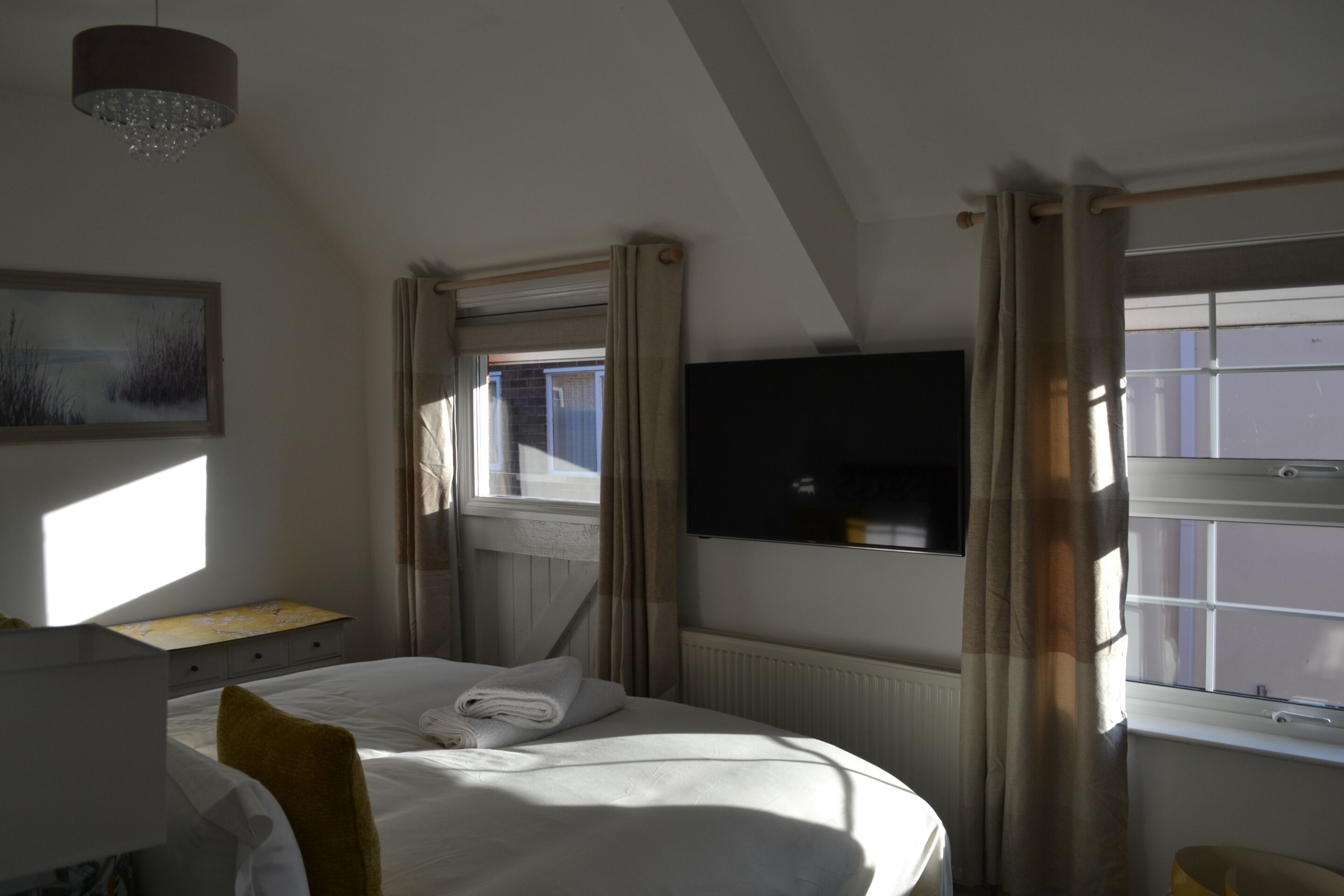 Et hotelværelse med en seng, et fladskærms-TV monteret på væggen, et vindue med gardiner og en lysekrone.