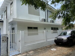 A white building with a gated entrance, air conditioning units, and a parked car. - Casa Vista (Anasco)