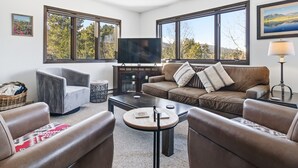 A modern living room with leather sofas, a coffee table, a TV, and large windows offering a view of trees and mountains. - Remodeled Nice Top-Floor Condo. Hot Tub. Fireplace (Breckenridge)