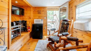 Un accogliente interno di una baita, con arredamento in legno, una televisione a parete, un frigorifero, un forno a microonde, una poltrona a dondolo in legno e una finestra con condizionatore.