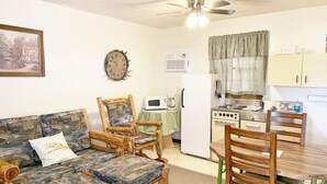 Un espace de vie compact comprenant un canapé, deux fauteuils, une table à manger, un coin cuisine et un ventilateur de plafond.