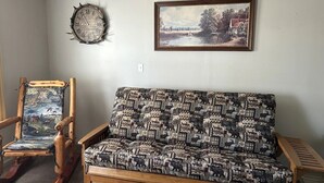 Un salon avec un fauteuil à bascule en bois, un canapé à motifs, une horloge murale et un tableau représentant un paysage.