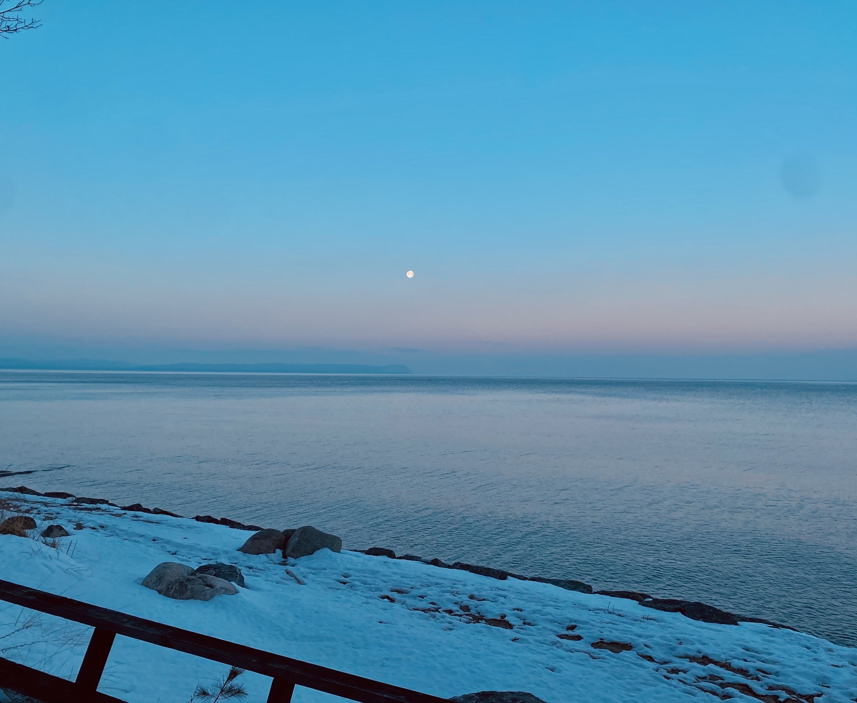 Un paisaje nevado con barandilla de madera, una masa de agua tranquila y una luna visible en el cielo.