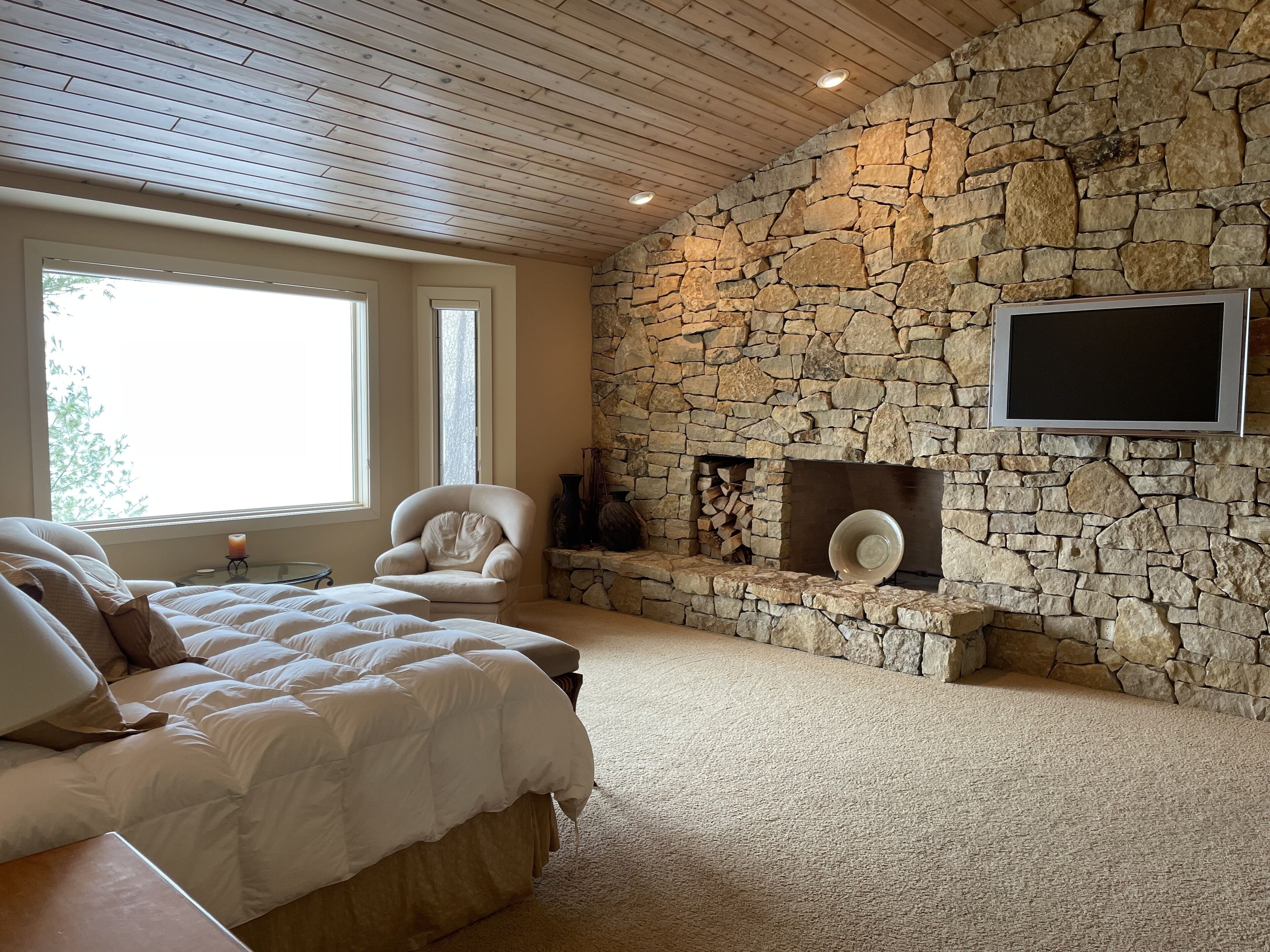 Un dormitorio con chimenea de piedra, una cama con ropa de cama blanca y un televisor montado en la pared.