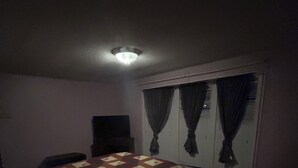 Un dormitorio tenuemente iluminado con una cama, una mesita, un televisor sobre un mueble bajo y una ventana con cortinas.