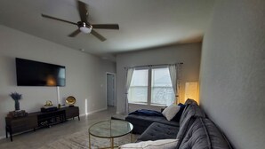 A modern living room with a flat-screen TV, a ceiling fan, a sofa, a coffee table, and a window with blinds. - New Modern home only 11 miles from Downtown and the Pearl Brewery! (Converse)