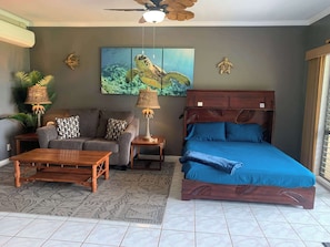 A room with a bed, sofa, and coffee table, a ceiling fan, and a painting of sea turtles.