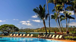 A pool with lounge chairs and palm trees, surrounded by a fence and a view of a mountain range.