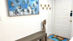 A hallway with a wooden bench, a door, a wall-mounted painting of palm trees, and a doormat with a pineapple design. - Minutes to Sanibel with Heated Pool, Tennis & Beach Gear (Fort Myers)